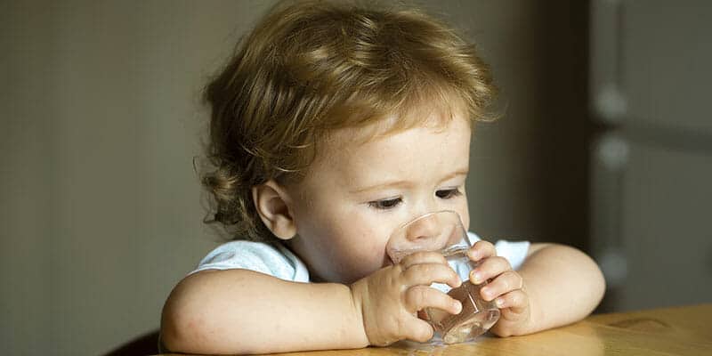 Kleines blondes Kind lehnt auf einem Tisch und trinkt Wasser