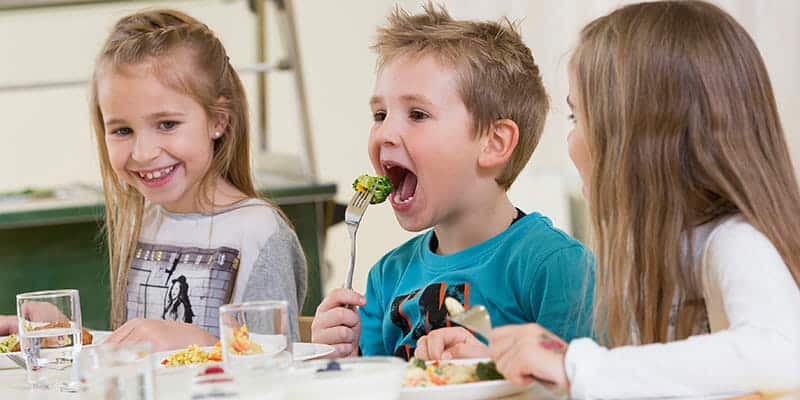 Kind isst begeistert Gemüse, zwei andere schauen lachend zu