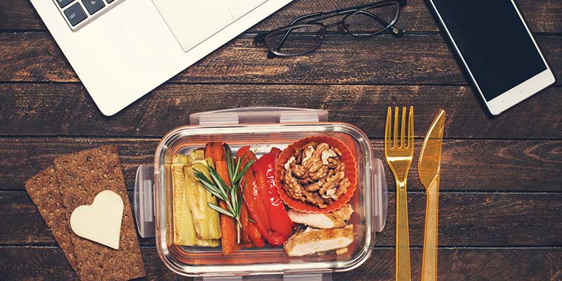 Auf einem Holztisch liegt eine Lunchbox mit gesundem Essen, eine Brille und ein Laptop